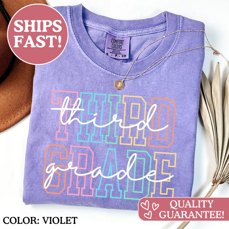 Puede incluir: Camiseta de color violeta con las palabras "third grade" en una fuente colorida y delineada. La camiseta tiene cuello redondo y un collar dorado. El texto "COLOR: VIOLET" est&aacute; en la parte inferior izquierda.
