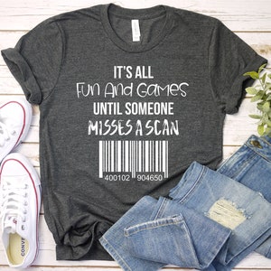 May include: Dark gray t-shirt with the text "It's All Fun And Games Until Someone Misses A Scan" in white. Below the text is a barcode and the numbers 400102 904650. The shirt is paired with white sneakers and blue jeans.