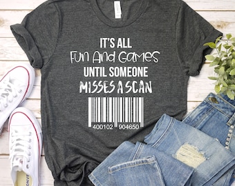 May include: Dark gray t-shirt with the text "It's All Fun And Games Until Someone Misses A Scan" in white. Below the text is a barcode and the numbers 400102 904650. The shirt is paired with white sneakers and blue jeans.