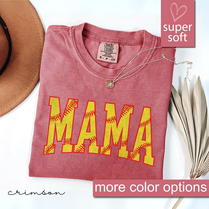 May include: Crimson red t-shirt with the word "MAMA" in yellow letters outlined in red. The letters are made up of softball stitches. The shirt is made of a soft, comfortable fabric.