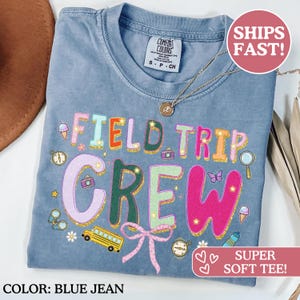 Puede incluir: Camiseta azul vaquero con las palabras "FIELD TRIP CREW" en letras coloridas y brillantes. La camiseta presenta ilustraciones de un autobús escolar, conos de helado, cámaras y una brújula. La camiseta está etiquetada como "SUPER SOFT TEE!"