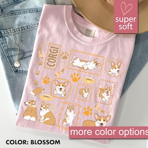 May include: A light pink t-shirt with a repeating pattern of Corgi dog illustrations in various poses, paw prints, and food bowls. The word "CORGI" is printed in a rectangular frame. The shirt is labeled "super soft" and "more color options".