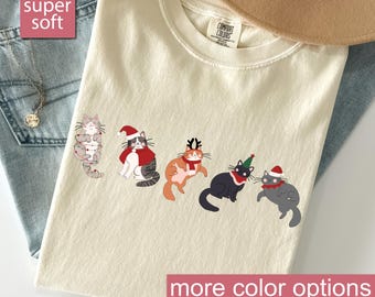 Puede incluir: Camiseta de color marfil con un diseño de gatos navideños. El gráfico incluye cinco gatos de dibujos animados con accesorios festivos. La camiseta tiene el texto "super soft" y "more color options".