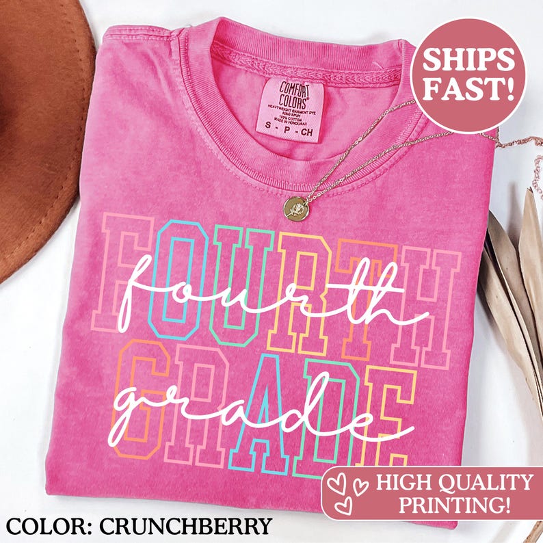 May include: A pink t-shirt in "Crunchberry" with the words "Fourth Grade" in a colourful design. The shirt has a crew neck and short sleeves. A gold necklace with a pendant is visible.