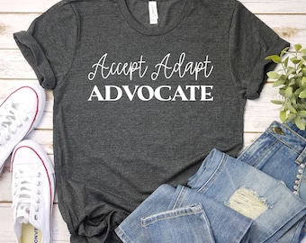 Może przedstawiać: Ciemnoszara koszulka z napisem "Accept Adapt Advocate" w białym skrypcie. Koszulka jest połączona z niebieskimi dżinsami i białymi trampkami. Koszulka jest wykonana z miękkiego materiału.