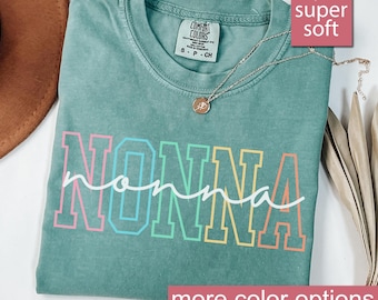 May include: A light green t-shirt with the word "NONNA" printed in rainbow colors. The shirt is made of a soft, comfortable fabric and has a relaxed fit.