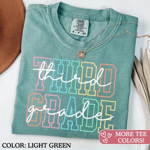 Puede incluir: Camiseta verde claro con las palabras "third grade" en un dise&ntilde;o de letras de bloque colorido. La camiseta tiene cuello redondo y mangas cortas. Un collar plateado con un peque&ntilde;o colgante est&aacute; sobre la camiseta.