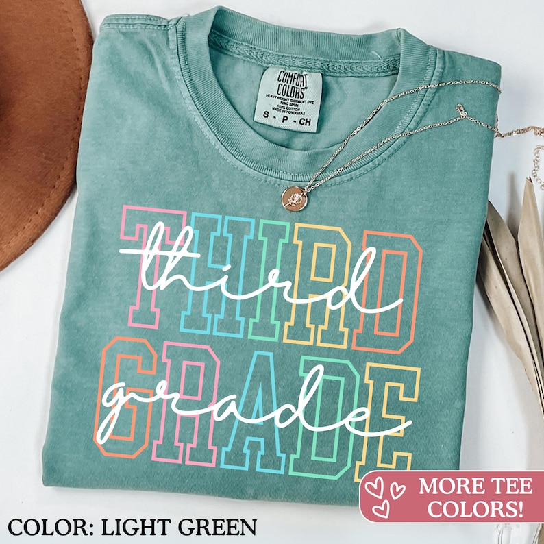 Puede incluir: Camiseta verde claro con las palabras "third grade" en un dise&ntilde;o de letras de bloque colorido. La camiseta tiene cuello redondo y mangas cortas. Un collar plateado con un peque&ntilde;o colgante est&aacute; sobre la camiseta.