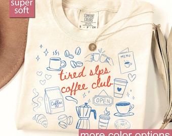 May include: Ivory-colored t-shirt with a "tired slps coffee club" design in red and blue. The shirt features coffee-related illustrations, including coffee cups, beans, and a coffee maker. A gold necklace with a pendant rests on the shirt.