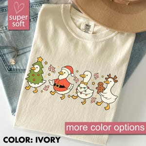 May include: Ivory t-shirt with a Christmas-themed design of cartoon ducks. The ducks are dressed as a Christmas tree, Santa, and a reindeer. The shirt also has the text "super soft" and "more color options".