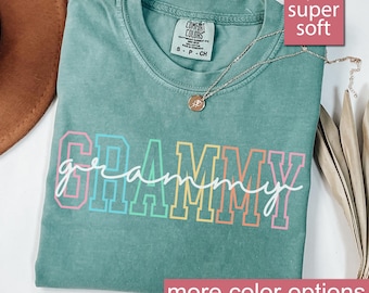 May include: A light green t-shirt with the word "GRAMMY" in rainbow colours. The word "grammy" is written in white script inside the rainbow letters. The shirt is made of a soft, comfortable fabric.