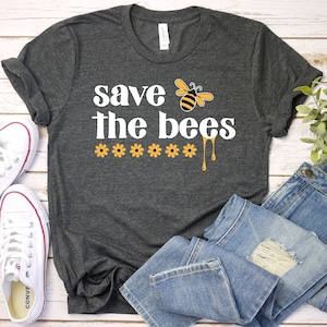 May include: A dark gray t-shirt with the text "save the bees" in white lettering. A cartoon bee with yellow and black stripes is above the text. Five yellow flowers are below the text with yellow dripping honey.