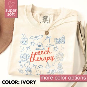 May include: Ivory-colored t-shirt with a graphic design featuring Halloween-themed doodles and the words "Speech Therapy" in red. The shirt also has the text "BOO!" and "more color options". A gold necklace is draped across the shirt.