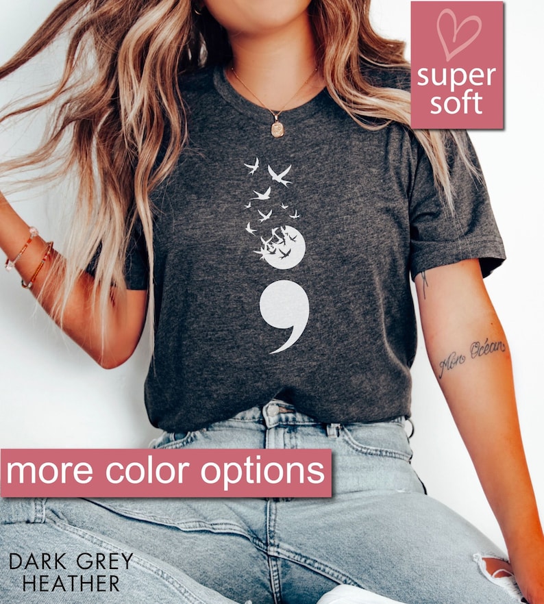 May include: A dark grey heather t-shirt with a white semicolon and a flock of birds flying away from it. The text "super soft" is in a pink heart shape above the shirt. The text "more color options" is in a pink rectangle below the shirt. The text "DARK GREY HEATHER" is below the rectangle.