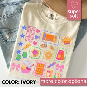 May include: Ivory t-shirt featuring a colourful baking-themed design with images of a microwave, cookies, a chef's hat, and a rolling pin. The text "NEEDS SALT" and "YEAST" are included. The shirt is labelled "super soft".