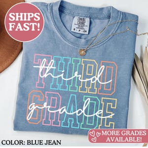 May include: Blue jean colored t-shirt with the words "Third Grade" in a colorful, block letter design. The word "third" is in a white cursive font. The shirt has a crew neck and a small tag at the top.