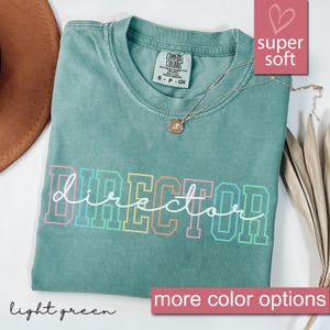 May include: A light green t-shirt with the word "DIRECTOR" written in rainbow colors. The shirt is made of a soft, comfortable fabric and has a relaxed fit.
