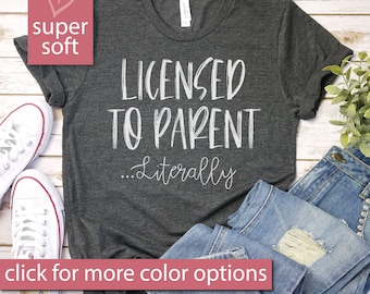 May include: A dark gray t-shirt with the text "LICENSED TO PARENT...Literally" in white lettering. The shirt is laid flat on a wooden surface with a pair of white sneakers and a pair of blue jeans.