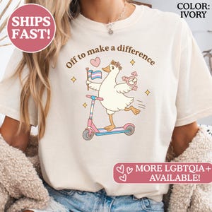 Puede incluir: Camiseta color marfil con un dibujo de un ganso en un patinete rosa y azul, sosteniendo una bandera transgénero y un símbolo de género. El texto "Off to make a difference" está sobre el ganso. La camiseta también tiene el texto "MORE LGBTQIA+ AVAILABLE!"