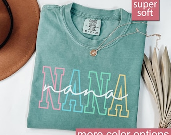 May include: A light green t-shirt with the word "NANA" in rainbow colors. The word "nana" is written in white script underneath. The shirt is made of a soft, comfortable fabric.