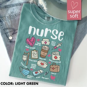 May include: Light green t-shirt with the word "nurse" and various medical-themed illustrations. The shirt features a stethoscope, coffee cup, syringe, and other medical symbols. The text "super soft" is in a pink heart-shaped box.