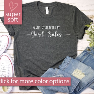 May include: A dark gray t-shirt with the text "Easily Distracted by Yard Sales" printed in white. The shirt is shown on a wooden surface with a pair of white sneakers and blue jeans.