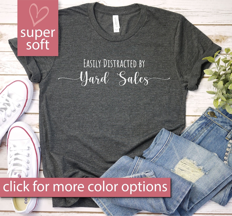 May include: A dark gray t-shirt with the text "Easily Distracted by Yard Sales" printed in white. The shirt is shown on a wooden surface with a pair of white sneakers and blue jeans.