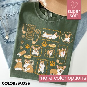 May include: Moss green t-shirt featuring a repeating pattern of Corgi dogs in various poses, paw prints, and dog toys. The word "CORGI" is printed in a rectangular frame. The shirt has a crew neck and short sleeves. The text "super soft" is in a pink box.