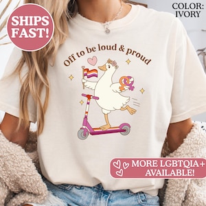 Puede incluir: Camiseta de color marfil con el texto "Off to be loud & proud". Un ganso de dibujos animados monta en un patinete rosa, sosteniendo una bandera del orgullo lésbico y un símbolo. La camiseta también tiene el texto "MORE LGBTQIA+ AVAILABLE!"