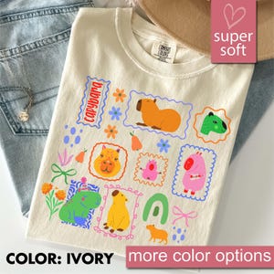 May include: Ivory-colored t-shirt featuring a colorful capybara design. The shirt has a crew neck and short sleeves. The design includes various capybara illustrations in different colors and poses, surrounded by floral and geometric elements. The text "capybara" is also present.
