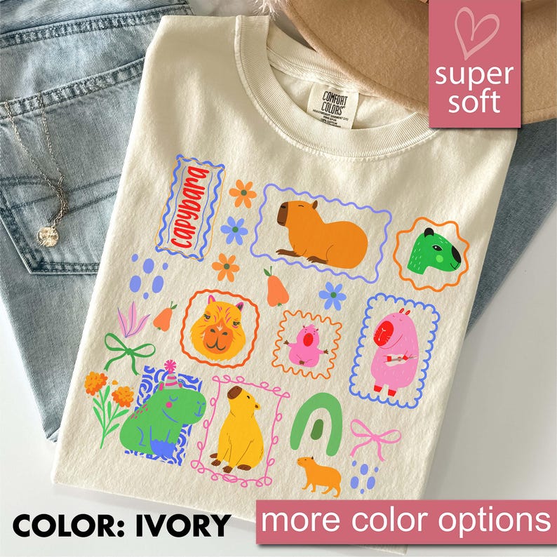 May include: Ivory-colored t-shirt featuring a colorful capybara design. The shirt has a crew neck and short sleeves. The design includes various capybara illustrations in different colors and poses, surrounded by floral and geometric elements. The text "capybara" is also present.