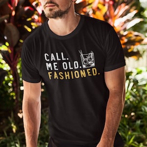 May include: A black t-shirt with the text "CALL ME OLD. FASHIONED." in white and gold lettering. A graphic of an old fashioned cocktail glass with ice and a straw is next to the text.