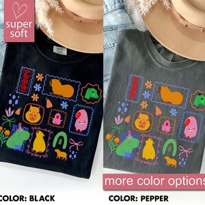 May include: Black and pepper-colored t-shirts with colorful capybara illustrations. The shirts feature various capybara designs within decorative frames, along with floral and geometric elements. The text "super soft" and "more color options" are also visible.