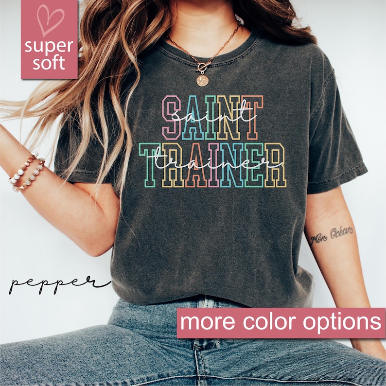 May include: A dark gray t-shirt with the text "SAINT TRAINER" in rainbow colored outlines. The shirt is made of a soft, comfortable material.