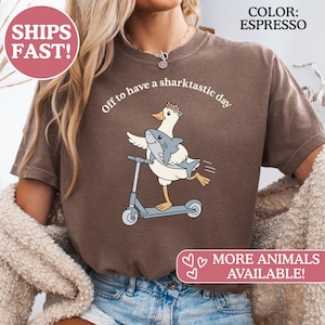 Op de afbeelding: Espresso-kleurig T-shirt met een cartoon eend op een scooter die een haai vasthoudt, met de tekst "Off to have a sharktastic day." Het shirt heeft ook de tekst "MORE ANIMALS AVAILABLE!"