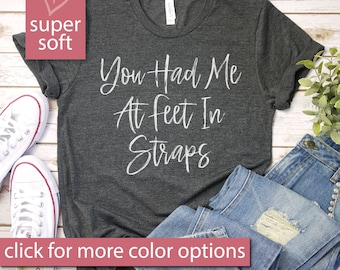 May include: A dark gray t-shirt with the text "You Had Me At Feet In Straps" printed in white. The shirt is laid flat on a wooden surface with a pair of white sneakers and a pair of blue jeans.