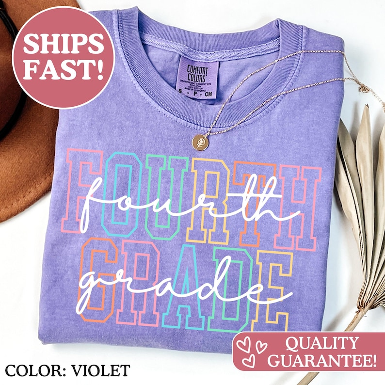 May include: A violet-coloured t-shirt with the words "FOURTH GRADE" in a colourful, outlined font. A gold necklace with a pendant rests on the shirt. The shirt is a Comfort Colors brand.
