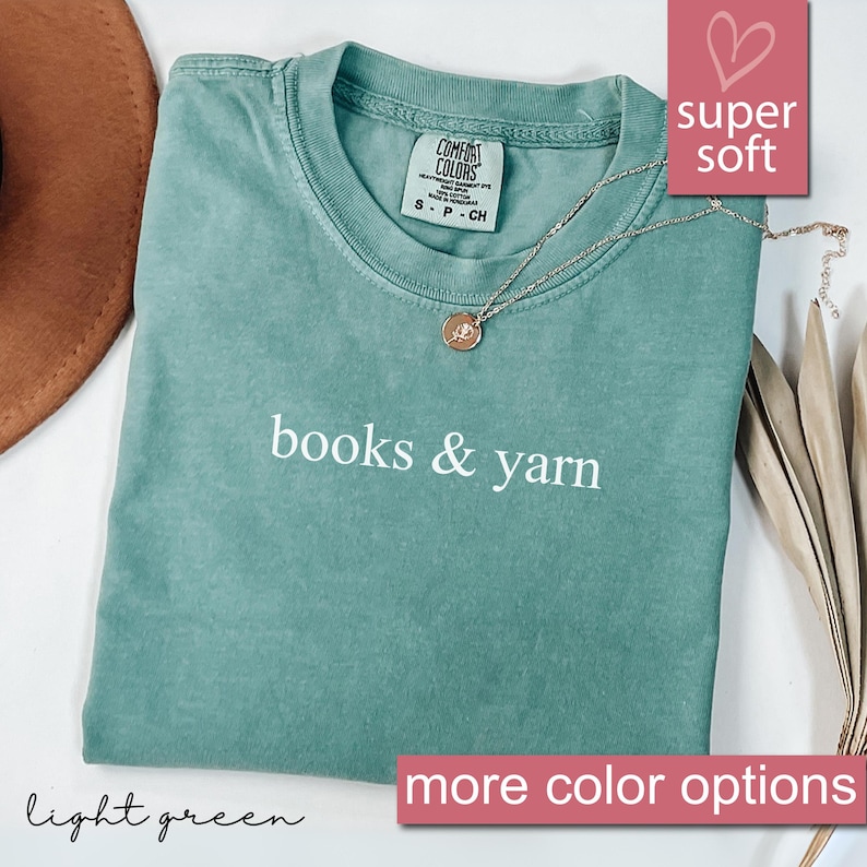 May include: A light green t-shirt with the words "books & yarn" in white. The shirt has a crew neck and short sleeves. A gold necklace with a pendant is draped on the shirt. The words "super soft" are in a pink box. The words "more color options" are in a pink box.