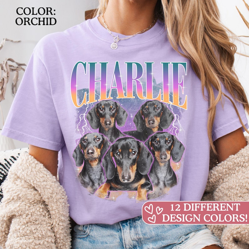 K&ouml;nnte beinhalten: Ein orchideenfarbenes T-Shirt mit einem Grafikdruck von vier schwarz-lohfarbenen Dackeln. Der Text CHARLIE ist in einer Verlaufsschrift &uuml;ber den Hunden gedruckt, mit Blitzakzenten und einem violetten Sternenhintergrund. Das T-Shirt hat kurze &Auml;rmel.