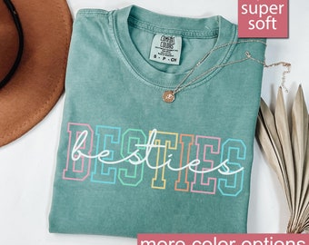 May include: A light green t-shirt with the word "besties" printed in rainbow colors. The shirt is made of a soft, comfortable fabric.