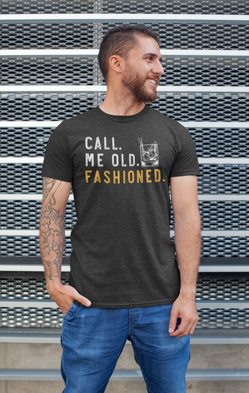 May include: A dark heather grey t-shirt with the text "CALL ME OLD. FASHIONED." in white with a graphic of an old fashioned cocktail in yellow.