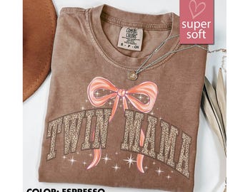 May include: Espresso-colored t-shirt with a large bow graphic and the text "TWIN MAMA" in a leopard print design. The shirt has a crew neck and short sleeves. A silver necklace with a pendant is visible.