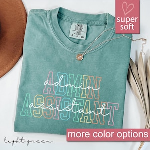 May include: A light green t-shirt with the words "ADMIN ASSISTANT" in a colorful, outlined font. The shirt has a crew neck and short sleeves. A silver necklace with a pendant is draped on the shirt. The words "super soft" and "more color options" are also visible.