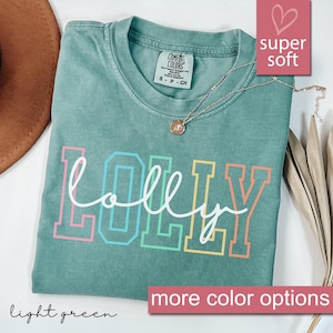 May include: A light green t-shirt with the word "LOLLY" in a colorful, outlined font. The shirt has a crew neck and short sleeves. A gold necklace with a pendant is draped on the shirt. The words "super soft" are in a pink box.