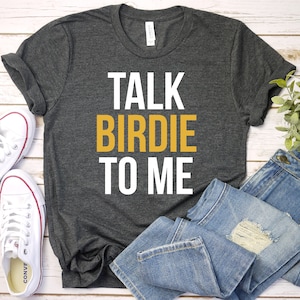 May include: A dark gray t-shirt with the text "TALK BIRDIE TO ME" in white and yellow lettering. The shirt is laid flat on a white wooden surface with a pair of white sneakers and blue jeans.