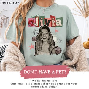 May include: A light green t-shirt with the name "Olivia" in a colorful, collage-style design. The shirt features a black and white image of a smiling person, disco balls, and star graphics. The text "DON'T HAVE A PET?" is also visible.