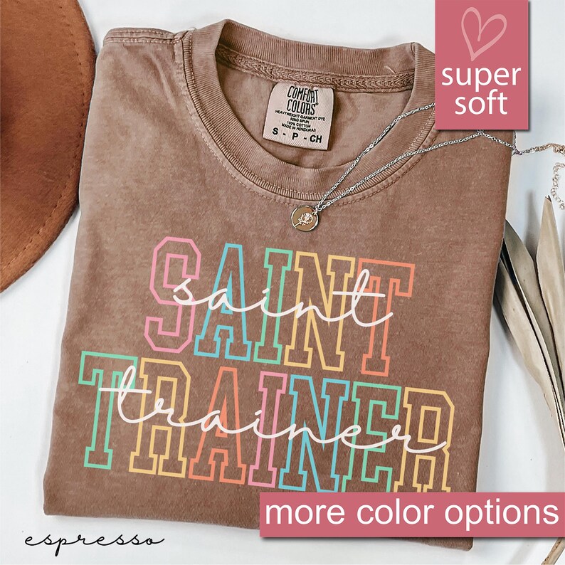 May include: A brown t-shirt with a distressed look featuring the text "ain't no saint trainer" in rainbow colored letters. The shirt is super soft and comfortable.