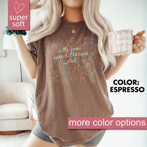 以下が含まれることがあります： エスプレッソ色のTシャツ。パステルカラーのガチョウのイラストと「silly goose speech therapy club」の文字がプリントされています。ゆったりとしたフィット感で、「super soft」と表示されています。