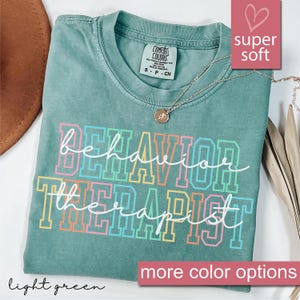 以下が含まれることがあります： レインボーカラーで「behavior therapist」と書かれたライトグリーンのTシャツ。柔らかく快適な生地で作られています。