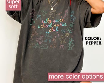 May include: Pepper-colored t-shirt with a whimsical design featuring cartoon geese and the text "silly goose school nurse club" in colorful lettering. The shirt also has the text "super soft" and "more color options".
